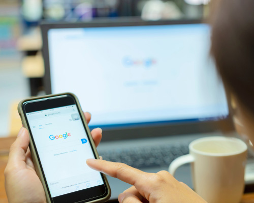 Hand mit Smartphone, auf dem die Google-Startseite zu sehen ist. Im Hintergrund ein aufgeklappter Laptop.